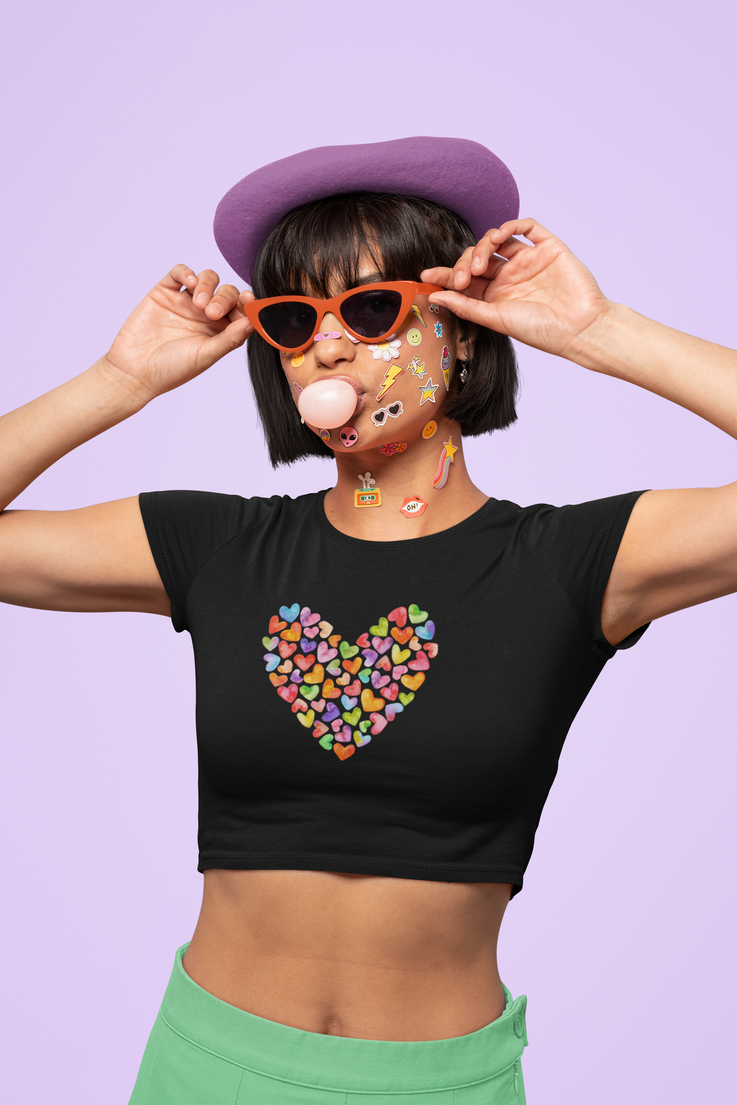 A person blows a bubblegum bubble, wearing orange sunglasses, a purple beret, and a black crop top with a colorful candy heart design. Their face is covered in playful stickers. The background is light purple.