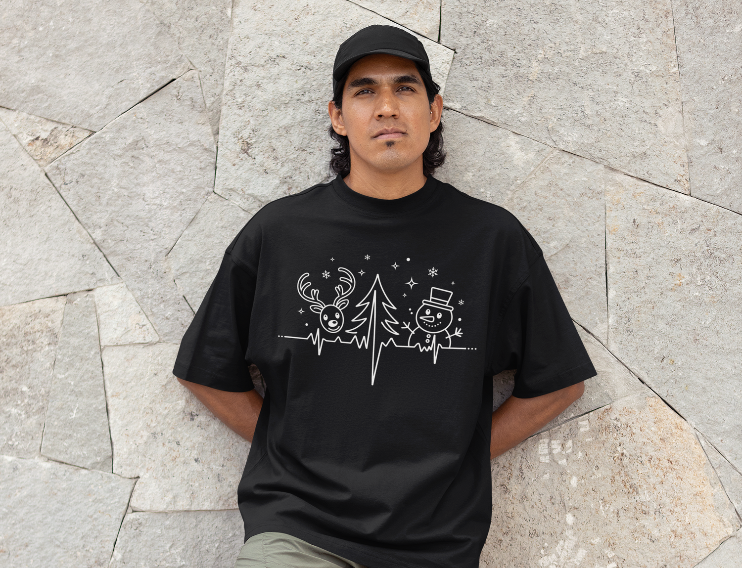A man in a black cap and black shirt stands against a stone wall. His shirt features a white graphic of a reindeer, a Christmas tree, and a snowman surrounded by stars and snowflakes.