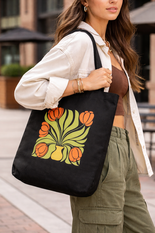 A woman in a beige shirt and green pants carries the Tulip Muse Tote by A Heartee Affair—a black cotton canvas bag with an orange tulip and green leaf design—while outdoors in an urban setting.