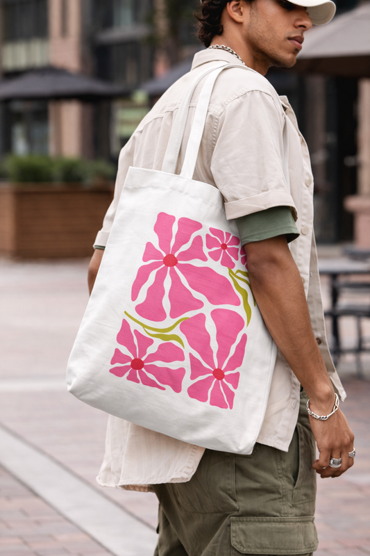 In an urban setting, a person in a beige shirt and cap carries the "Pink Petal Tote" by A Heartee Affair—an eco-friendly canvas bag with a vibrant pink and green floral design.