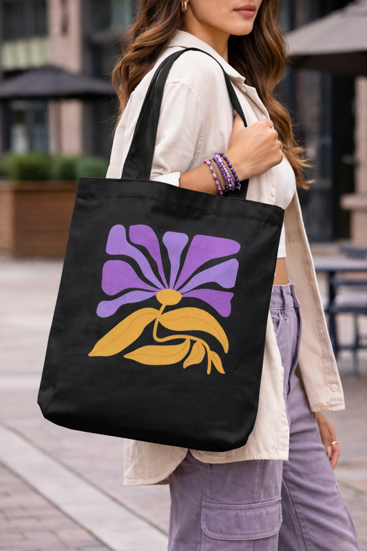 Outdoors, a woman carries the Violet Muse Tote by A Heartee Affair—an eco-friendly cotton canvas bag with a purple and yellow floral print. She pairs it with a light jacket, white top, purple pants, and stacked beaded bracelets.