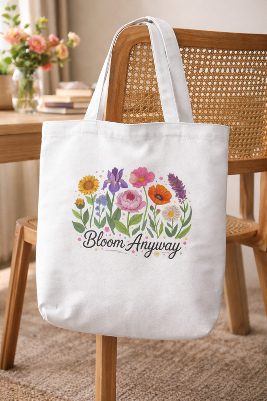 The Bloom Era Tote by A Heartee Affair, a white cotton canvas bag with colorful illustrated flowers and "Bloom Anyway" text, rests on a woven chair with a vase of flowers and table in the background.