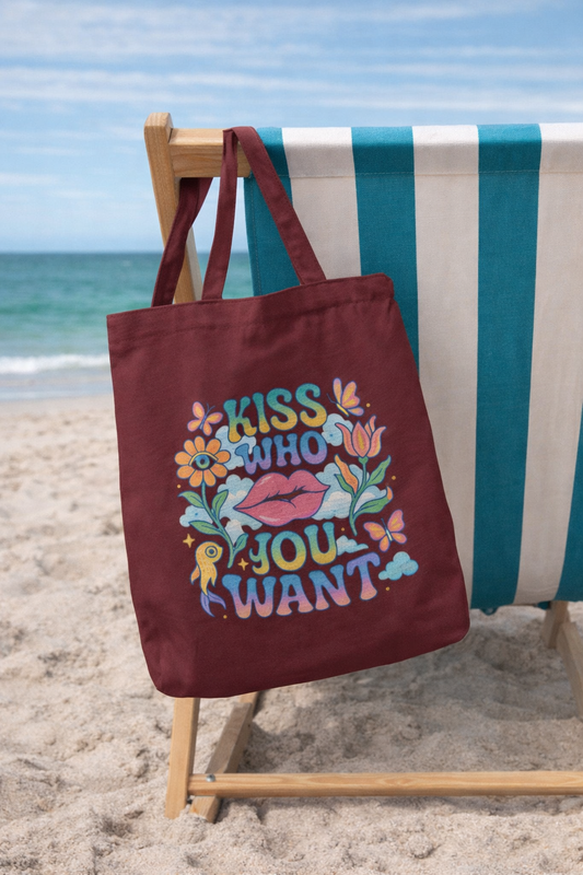 A woman in a white dress and sun hat holds the Lips and Liberation Tote by A Heartee Affair, an eco-friendly cotton canvas bag featuring colorful lips, flowers, and “Kiss Who You Want.” Palm trees and cars are in the background.