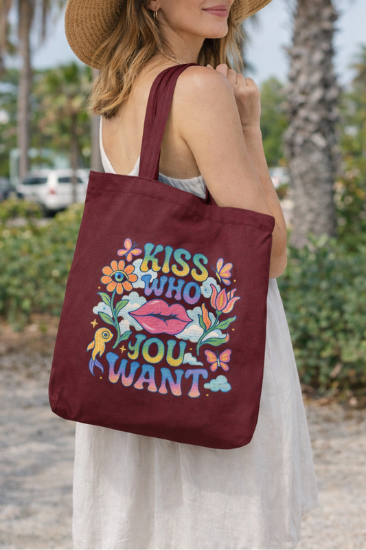 A woman in a white dress and sun hat holds the Lips and Liberation Tote by A Heartee Affair, an eco-friendly cotton canvas bag featuring colorful lips, flowers, and “Kiss Who You Want.” Palm trees and cars are in the background.