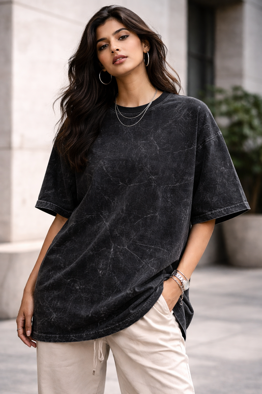 A woman with long dark hair stands outdoors near a modern building, confidently wearing the A Heartee Affair Unisex Acid Washed Oversized Tee, light beige pants, hoop earrings, and layered necklaces.