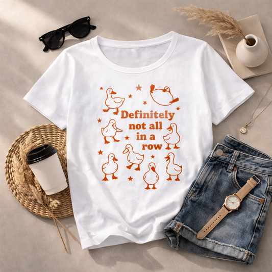 The Not In a Row Classic T-Shirt by A Heartee Affair is a white, 100% cotton tee featuring orange cartoon ducks and “Definitely not all in a row,” surrounded by stars, styled with denim shorts, sunglasses, jewelry, and coffee.