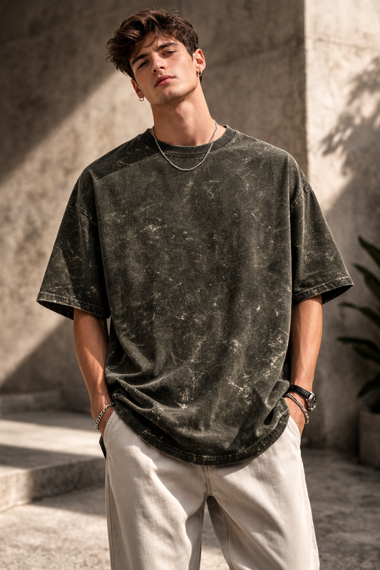 A young man stands outdoors with his hands in his pockets, wearing the A Heartee Affair Unisex Acid Washed Oversized Tee and light pants, looking confidently at the camera against a concrete background in natural sunlight.