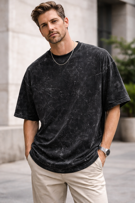 A woman with long dark hair stands outdoors near a modern building, confidently wearing the A Heartee Affair Unisex Acid Washed Oversized Tee, light beige pants, hoop earrings, and layered necklaces.