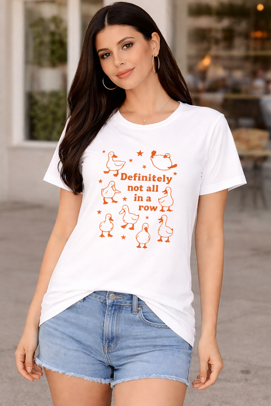 The Not In a Row Classic T-Shirt by A Heartee Affair is a white, 100% cotton tee featuring orange cartoon ducks and “Definitely not all in a row,” surrounded by stars, styled with denim shorts, sunglasses, jewelry, and coffee.