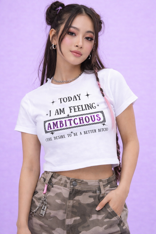 A young woman with long braids wears the Ambitchous Baby Tee by A Heartee Affair, featuring “Today I am feeling ambitchous (the desire to be a better bitch)” and camo pants against a light purple background—classic Gen Z style.