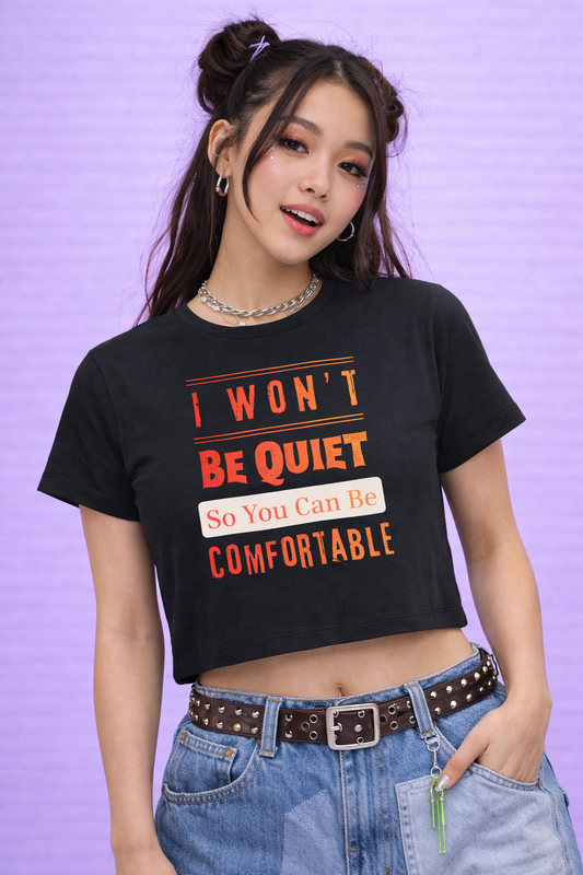 A young woman with styled hair and hoop earrings models the Unmuted Baby Tee by A Heartee Affair, featuring "I WONT BE QUIET So You Can Be COMFORTABLE." She poses against a light purple textured background—bold Gen Z style.