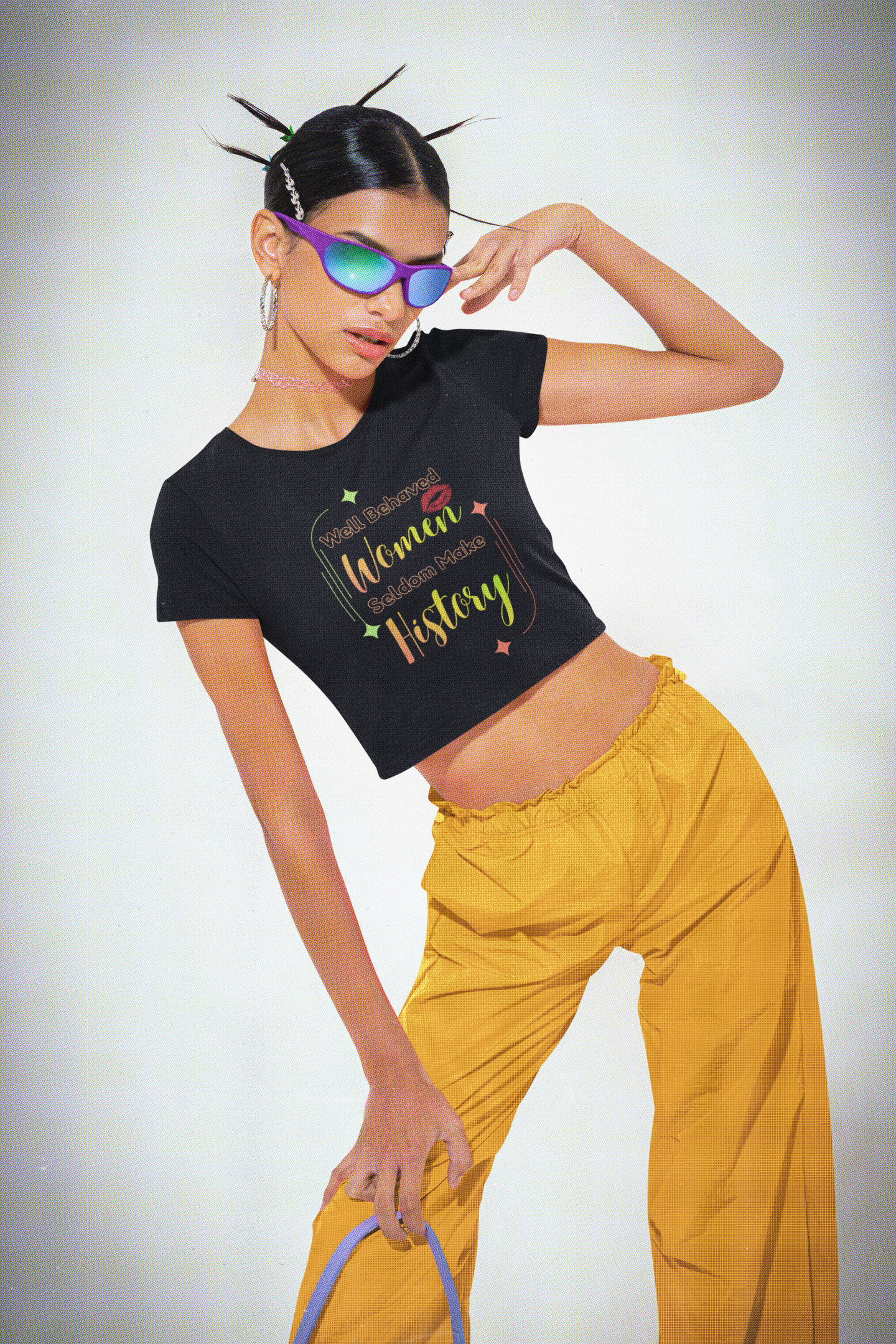 A woman poses confidently in the Bold Her Baby Tee by A Heartee Affair, featuring “Well Behaved Women Seldom Make History”—a cotton spandex cropped essential for any Gen Z wardrobe. She pairs it with sunglasses and yellow pants against a plain background.