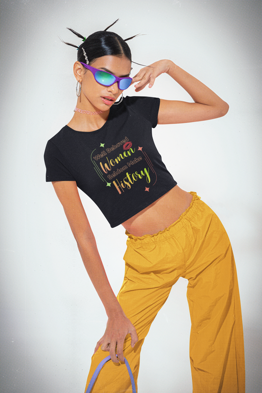 A woman poses confidently in the Bold Her Baby Tee by A Heartee Affair, featuring “Well Behaved Women Seldom Make History”—a cotton spandex cropped essential for any Gen Z wardrobe. She pairs it with sunglasses and yellow pants against a plain background.